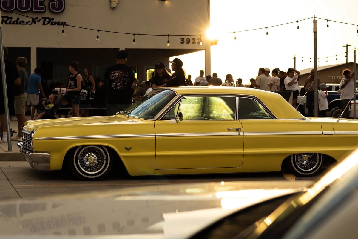 A vintage golden-yellow sedan from the 1960s parked at a car show with crowds and string lights in the background