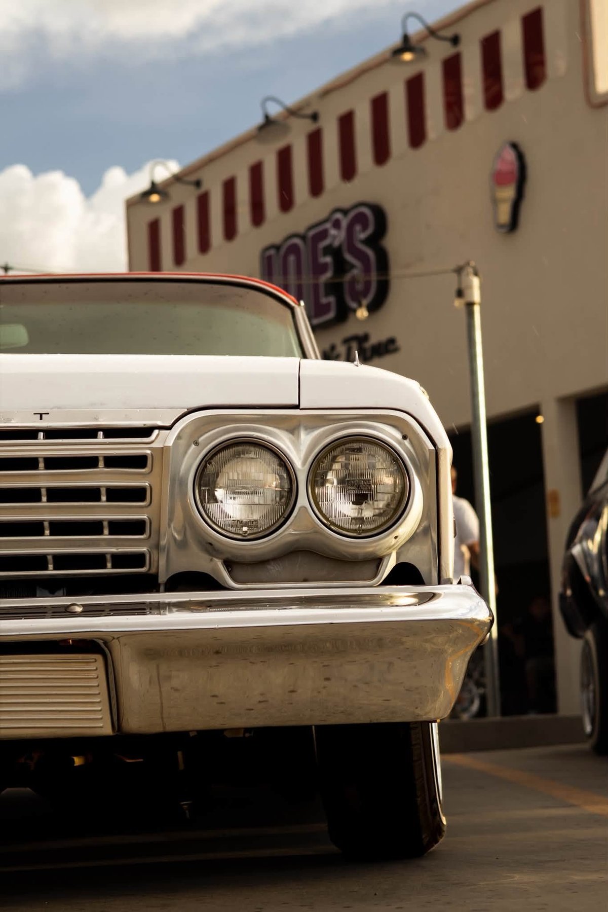 Close-up of a vintage white car with dual round headlights parked in front of a beige building with a neon sign