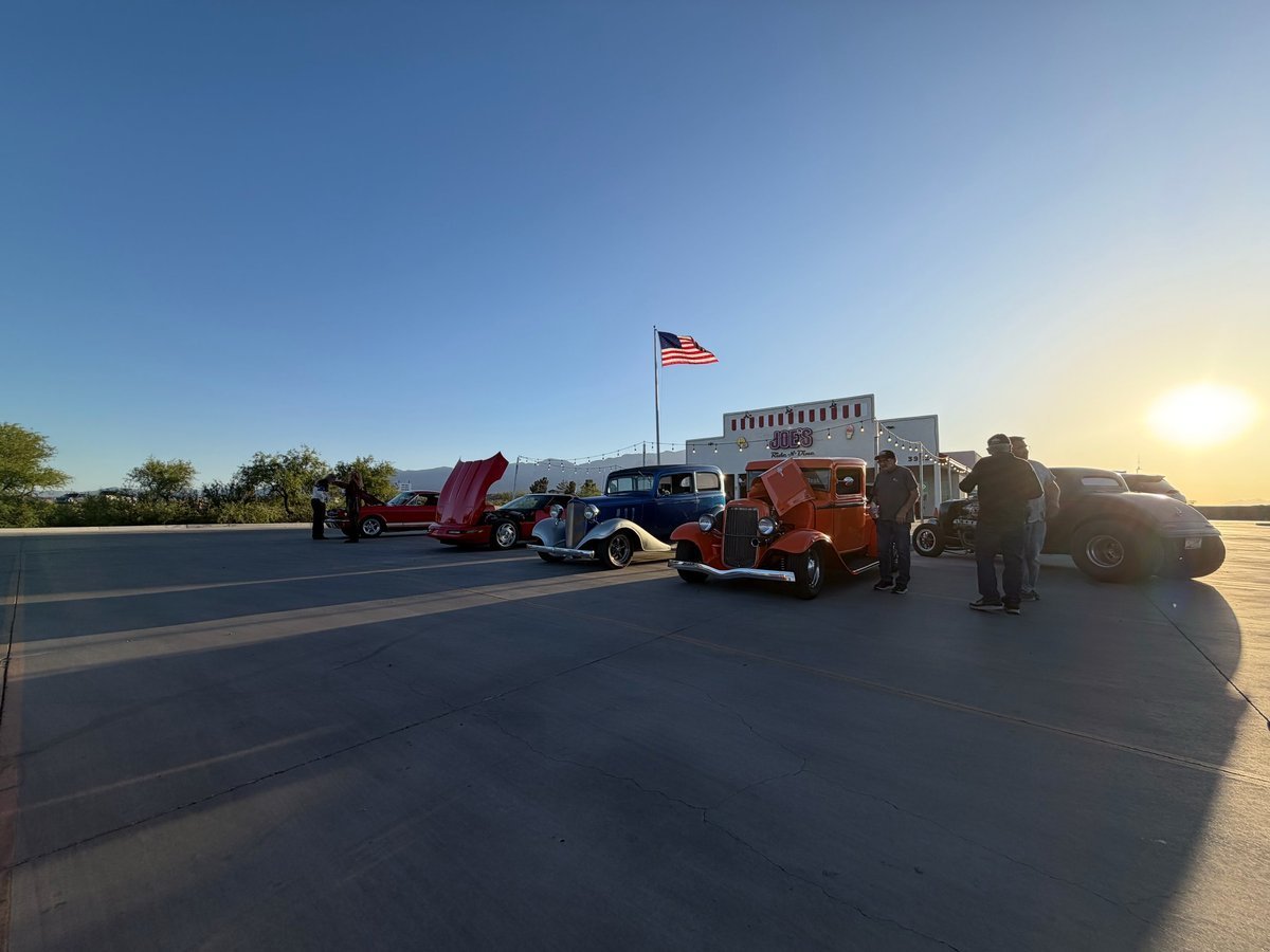 Vintage cars and trucks displayed on a desert road with an American flag flying overhead during sunset